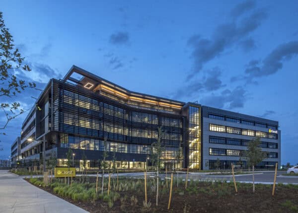 Office building illuminated at dusk, its modern glass-and-metal facade with horizontal sunshades glowing from interior lights, surrounded by young trees, landscaping, and a paved entrance road.

Text in image: "Kiewit" (appears on a yellow ground sign and on the building facade).