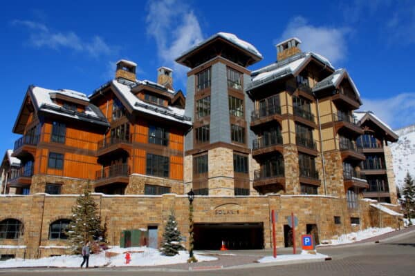 Multi-story timber-and-stone ski-resort building stands with snow-topped gabled roofs beneath a clear blue sky; a pedestrian walks along the snowy sidewalk. 
Text visible: "SOLARIS", "P"