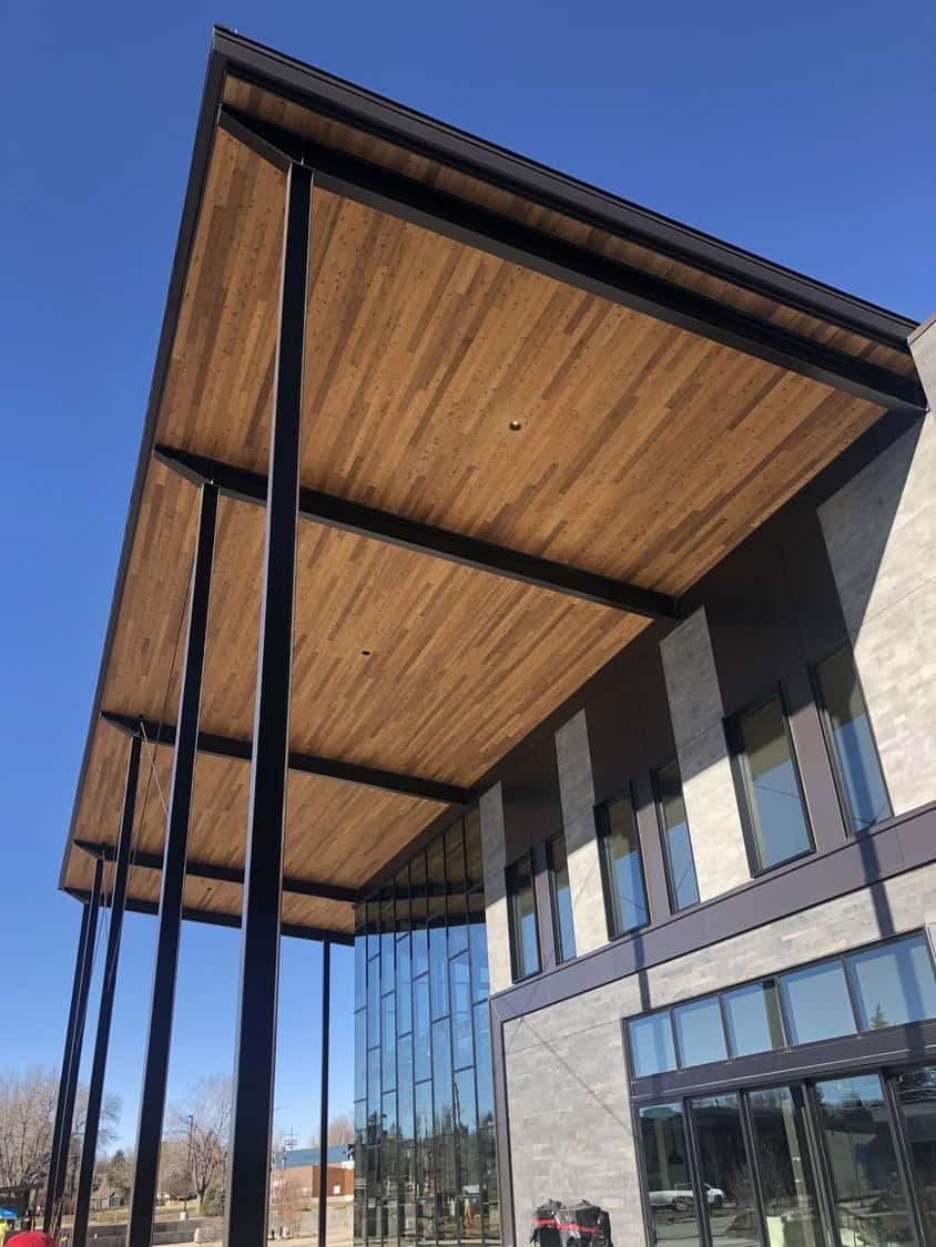 Large wooden canopy extends over the entrance, supported by tall black steel columns, sheltering a modern glass-and-stone commercial building set against a clear blue sky.