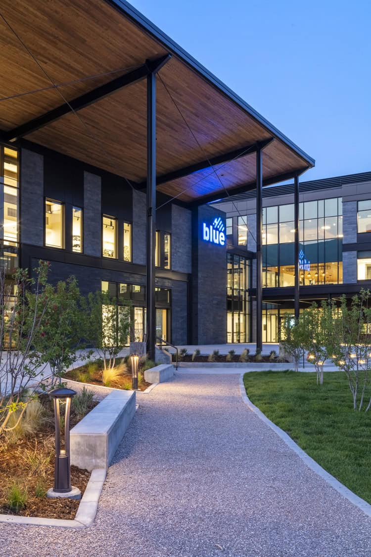 Modern office building entrance displays illuminated sign reading "blue", with a lit gravel pathway, landscaped courtyard, and overhanging wooden canopy at dusk.