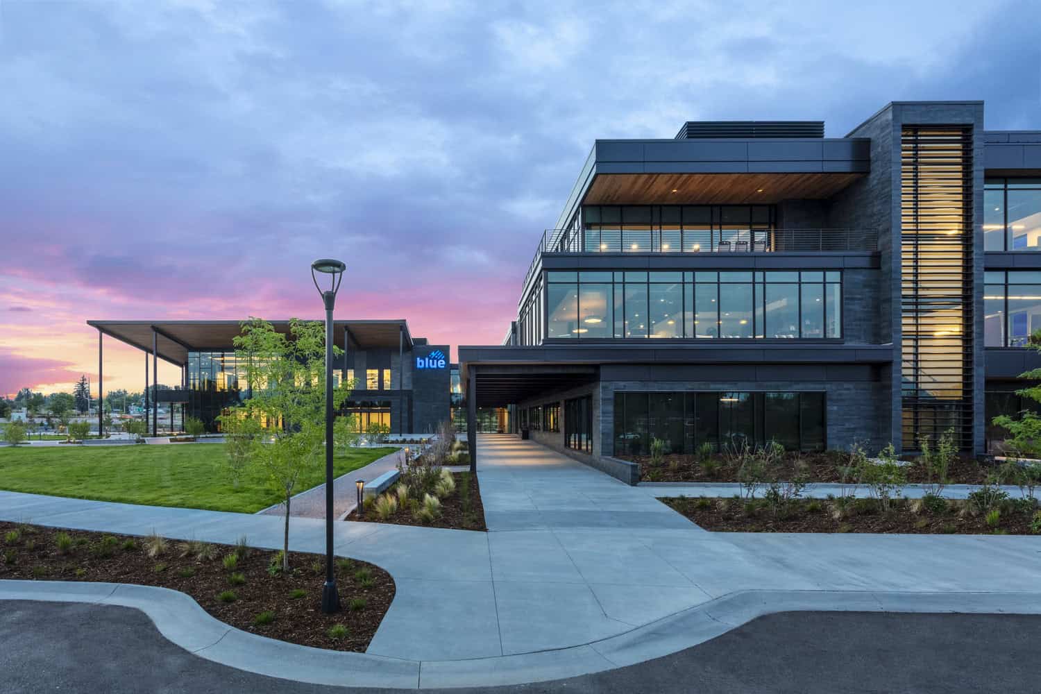 Modern glass-and-stone office building — illuminated, with a covered walkway leading inward — set on a landscaped campus at dusk beneath a pink-purple sunset sky. Sign reads "blue".