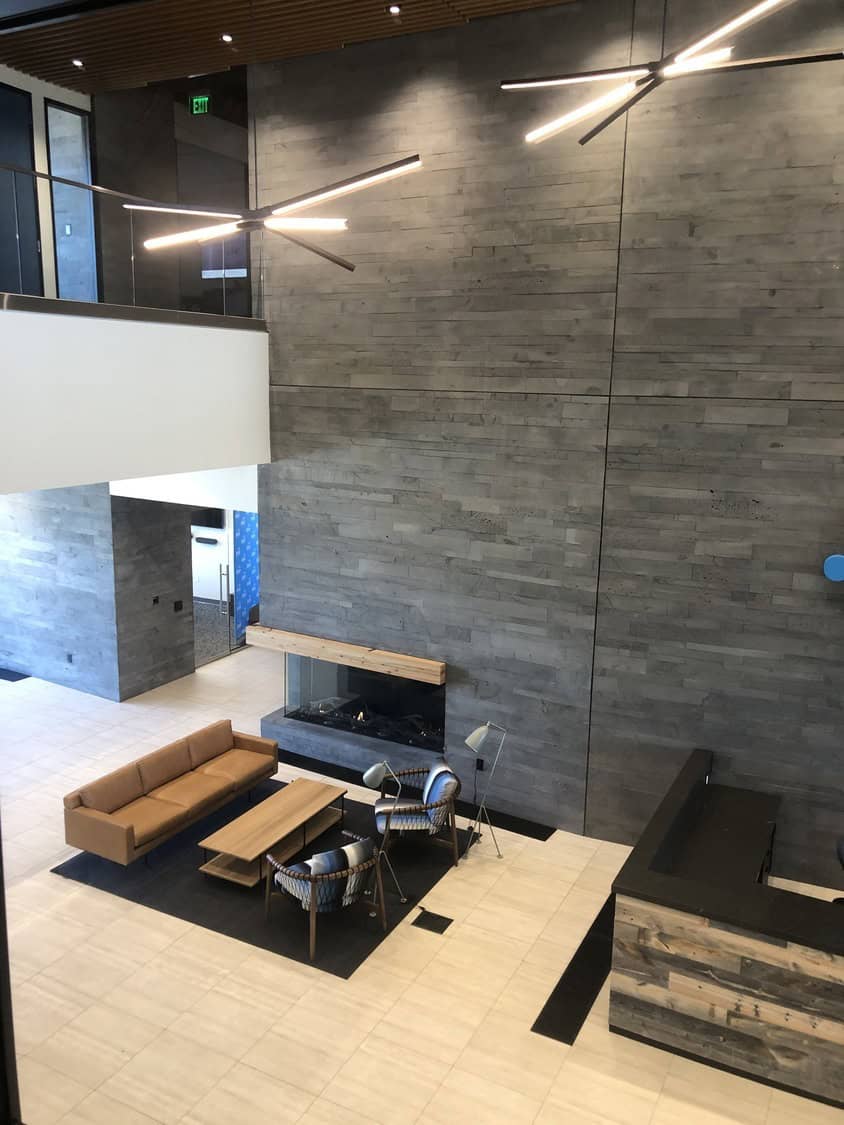 Seating area: tan sofa and two woven armchairs arranged around a wooden coffee table facing a recessed fireplace on a dark rug in modern lobby with gray wood wall. EXIT