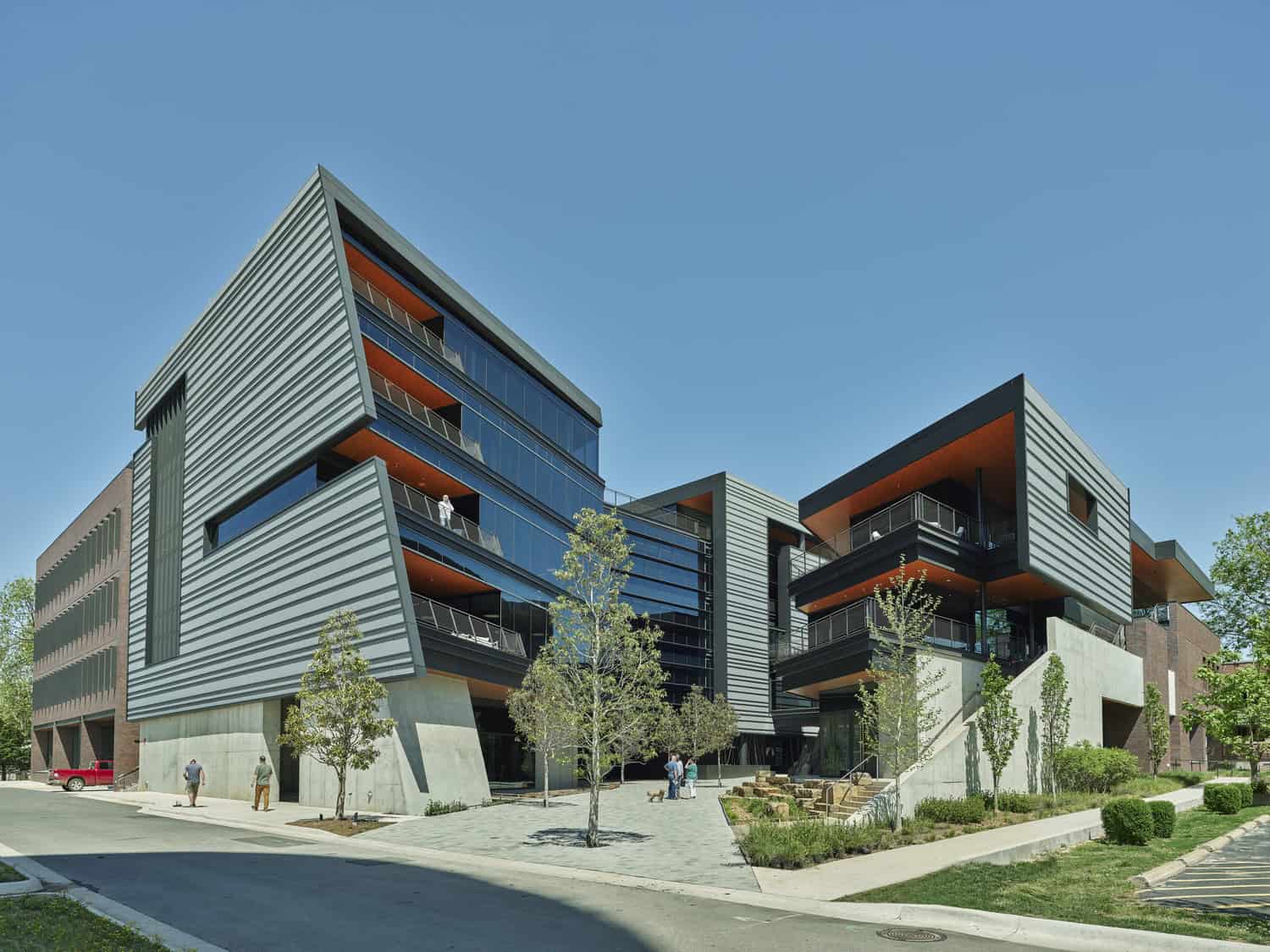 Modern angular office complex juts forward with layered metal-clad volumes and glass balconies; people walk in a tree-lined courtyard beside concrete stairs and a street under a clear blue sky.
