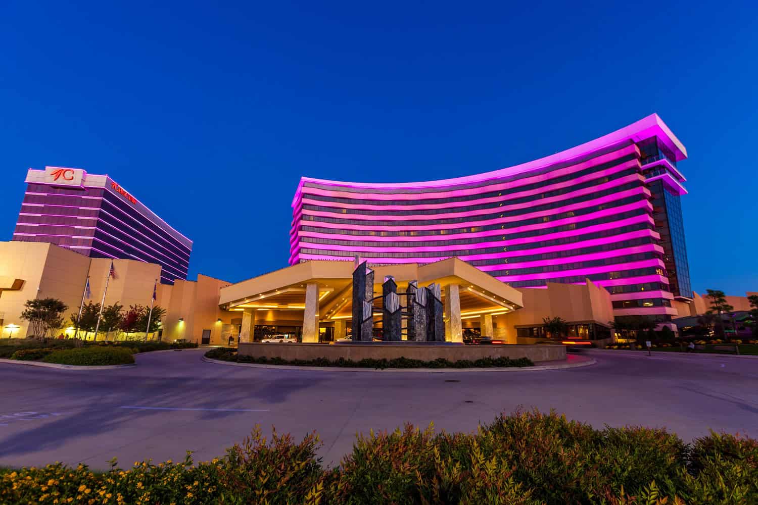 Curved hotel tower glows in magenta banded lighting, rises behind a lit porte-cochère sheltering vehicles around a central sculptural fountain, set against a deep twilight sky with landscaped driveway foreground.