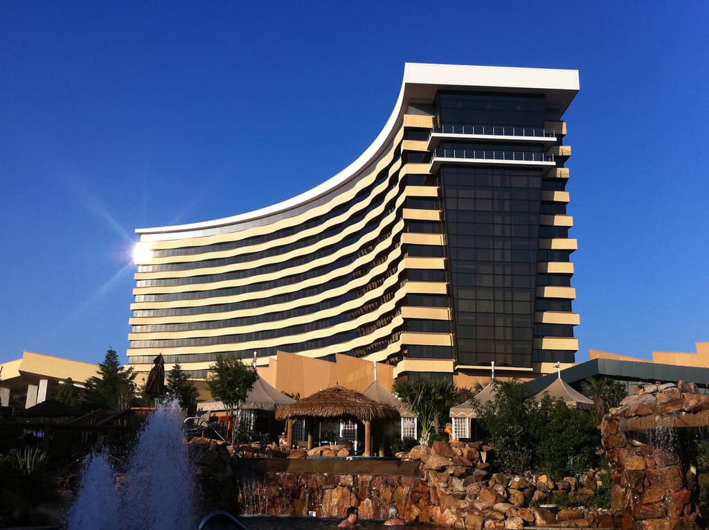 Curved multi-story hotel rises with staggered balconies, sunlight glinting off glass; a fountain spurts in the foreground amid rocky landscaping, thatched cabanas, and pools under a clear blue sky.