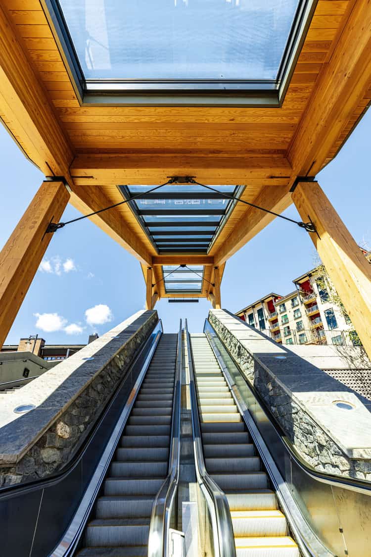 A pair of outdoor escalators ascend beneath a wooden pergola with glass skylights, framed by stone sidewalls, leading upward toward a clear blue sky and nearby apartment buildings.