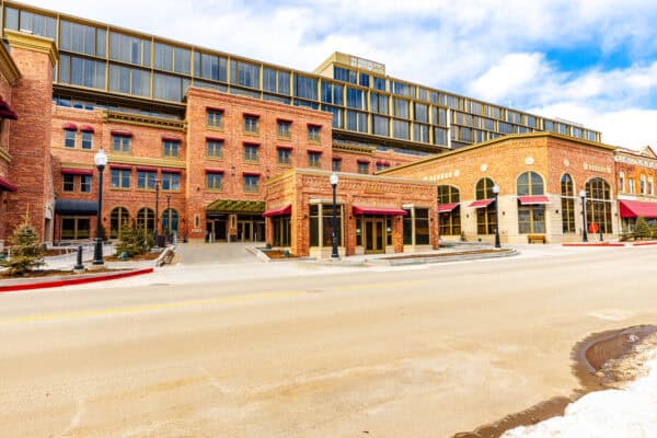 Brick hotel complex rises with red-awned storefronts and lamp posts, backed by a modern glass upper addition; an empty, slightly snow-edged street runs in the foreground under a cloudy sky. No legible text.