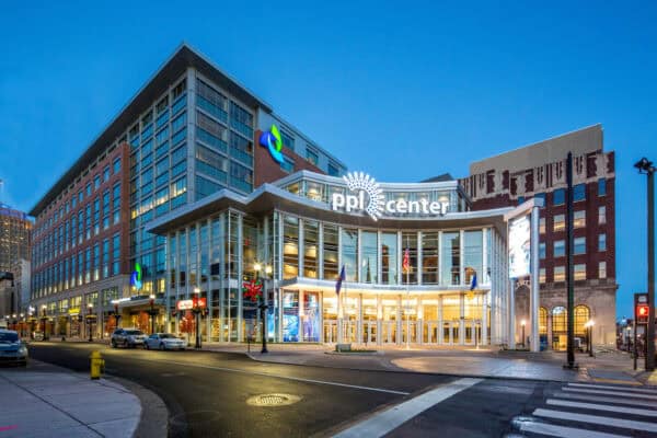 PPL Center building glows with an illuminated "ppl center" sign; its glass-front entrance (TIM HORTONS GATE) is warmly lit, flags flying, storefronts including "Tim Hortons" line a downtown street at dusk.