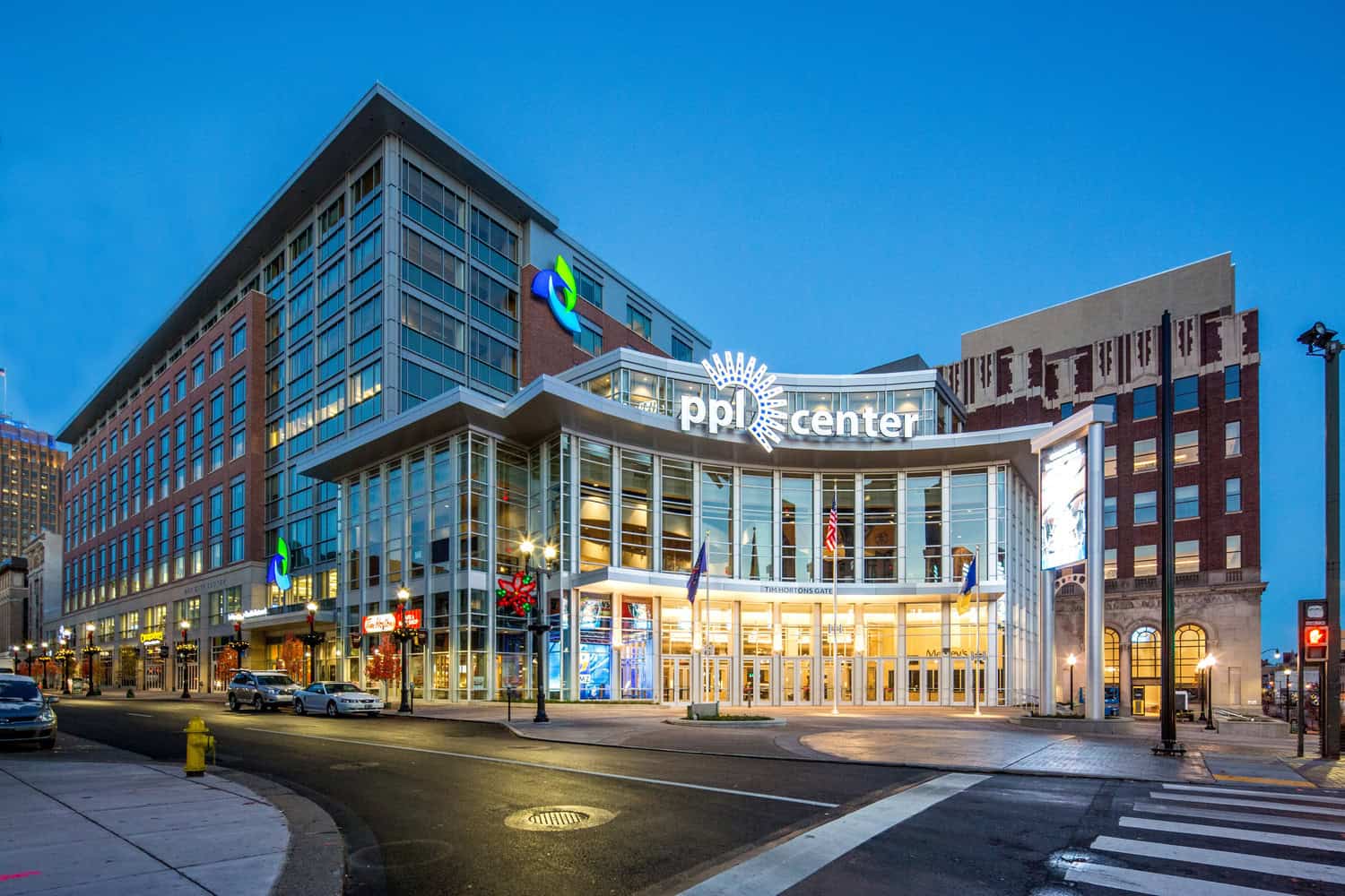 PPL Center building glows with an illuminated "ppl center" sign; its glass-front entrance (TIM HORTONS GATE) is warmly lit, flags flying, storefronts including "Tim Hortons" line a downtown street at dusk.