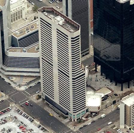 Tall rectangular office tower anchors a downtown intersection, rising above adjacent skyscrapers and plaza areas, with surrounding streets, moving and parked cars, and a nearby surface parking lot.