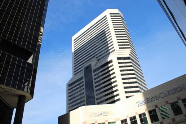 A tall white-and-glass skyscraper rises upward, casting shadows between neighboring dark reflective towers against a clear blue sky. Visible text: "TRU FIT ATHLETIC CLUBS" and "THE RITZ-CARLTON".