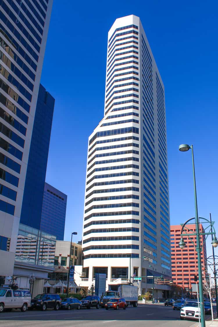 A tall white-striped office tower rises above a busy downtown street, flanked by other skyscrapers, parked cars and trucks, with streetlamps and pedestrians under a clear blue sky.