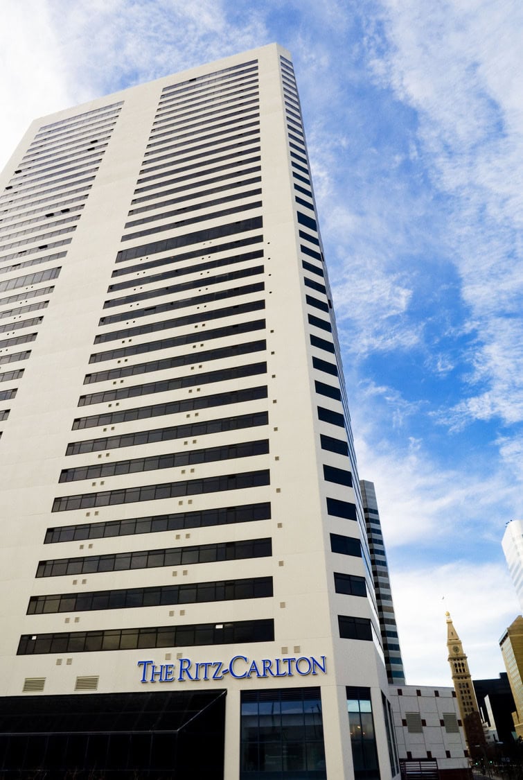 A tall white hotel tower rises skyward, dominating a downtown block with neighboring office buildings and a distant clock tower under a bright blue, streaked sky. Text: "The Ritz-Carlton."