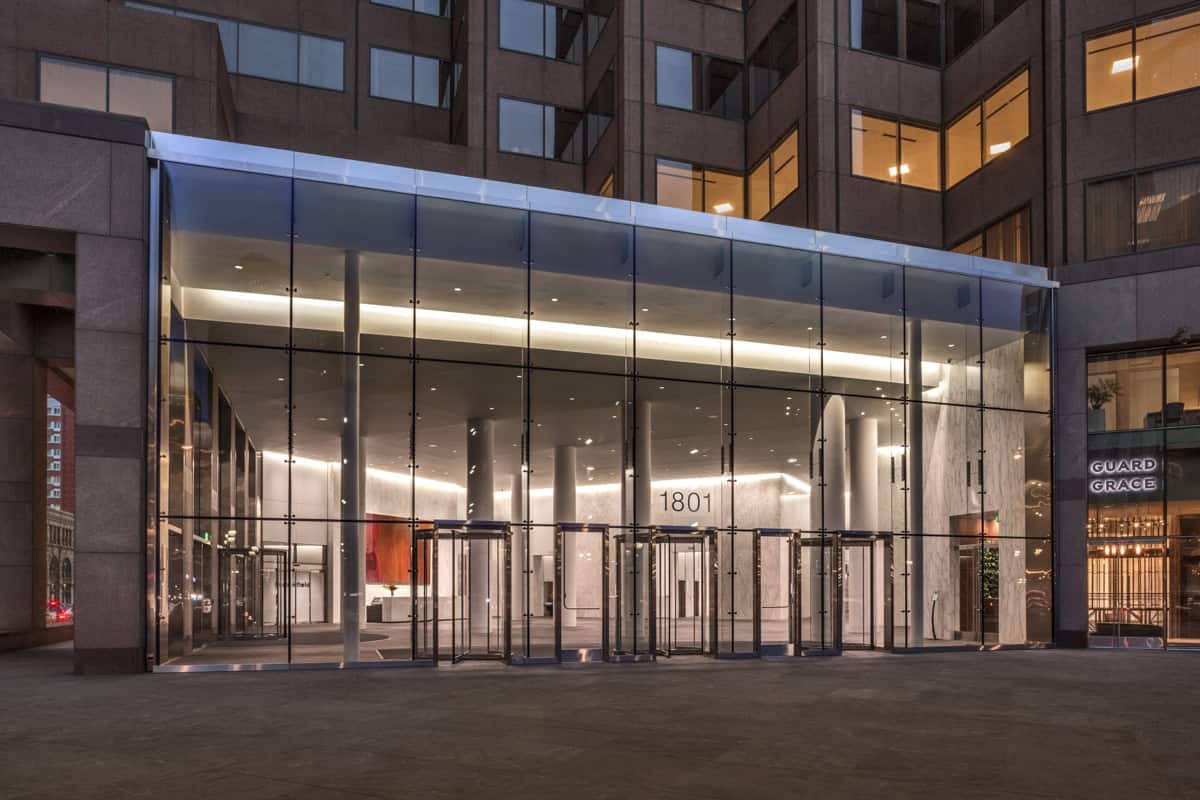 Glass office entrance with revolving doors and lit marble lobby, framed by high-rise facade and plaza at dusk; building number "1801" visible above doors; storefront sign reads "GUARD GRACE".