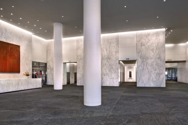 A tall white column stands centered; it anchors a marble-walled office lobby with a marble reception desk, abstract orange painting, elevators and doorways labeled "2-13", "15-26", "GARAGE" and a green "EXIT" sign.