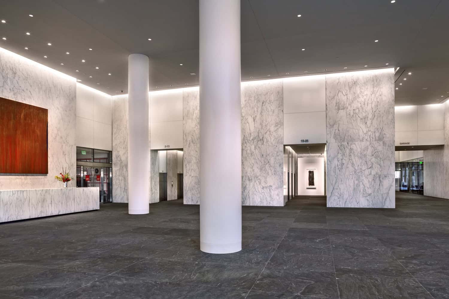 A tall white column stands centered; it anchors a marble-walled office lobby with a marble reception desk, abstract orange painting, elevators and doorways labeled "2-13", "15-26", "GARAGE" and a green "EXIT" sign.
