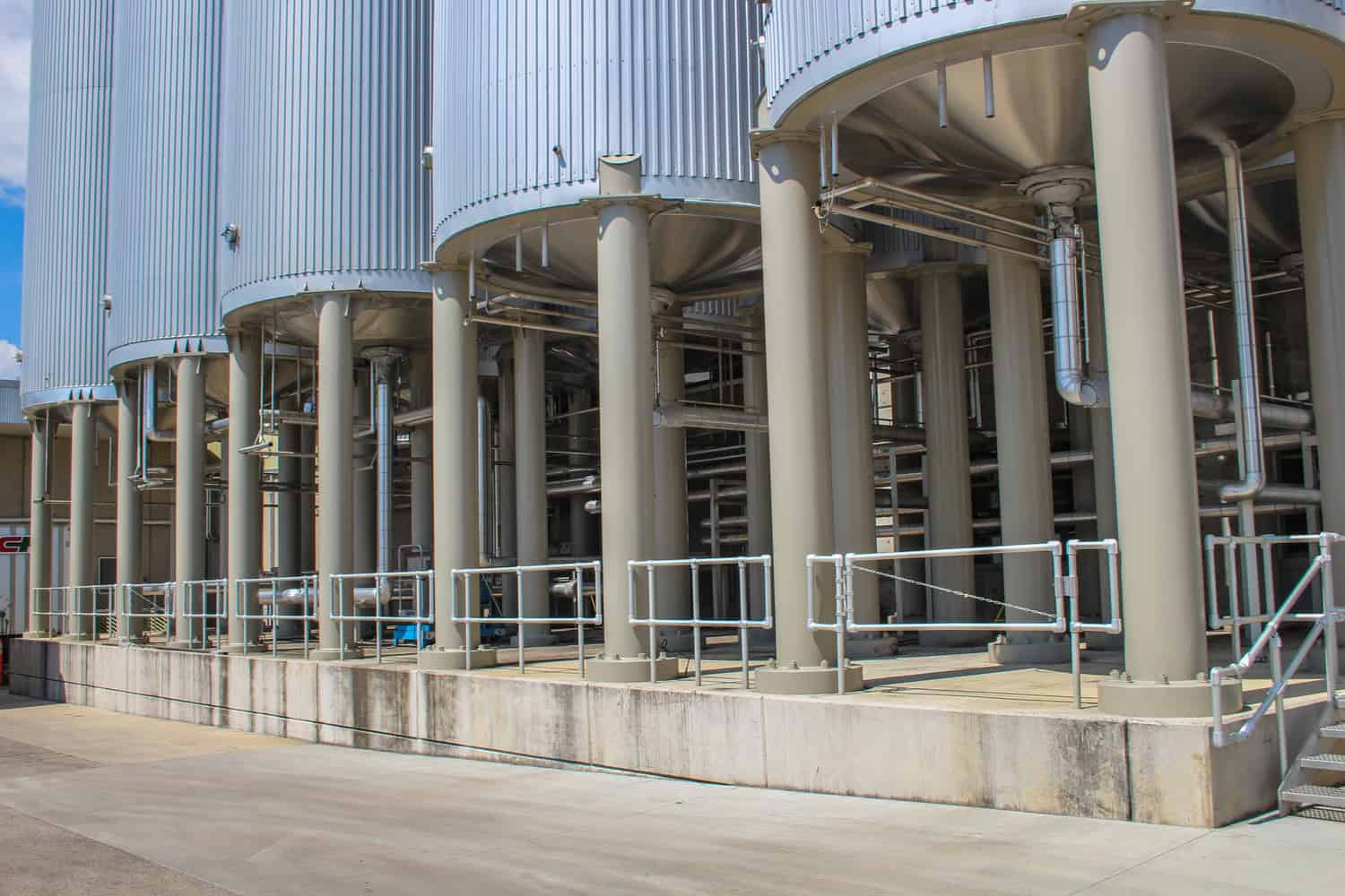 Several large corrugated metal storage silos stand on beige cylindrical supports, connected by stainless-steel pipes and walkways, above a concrete loading platform at an outdoor industrial processing facility.