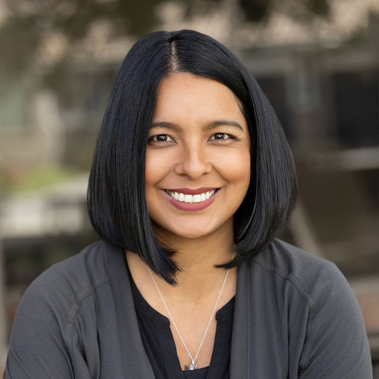 Smiling woman with straight, shoulder-length black hair wearing a dark cardigan and necklace smiles directly at the camera against a blurred outdoor background.