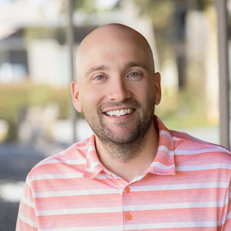 Bald man in a pink-and-white striped polo smiles at the viewer; short beard and light green eyes, seated outdoors with a soft, blurred background and warm natural light.