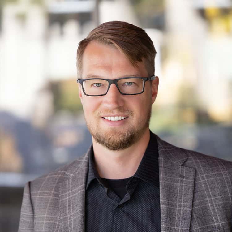 A smiling man with short hair, a trimmed beard and rectangular glasses faces the camera, wearing a plaid blazer over a dark shirt against a softly blurred outdoor/urban background.