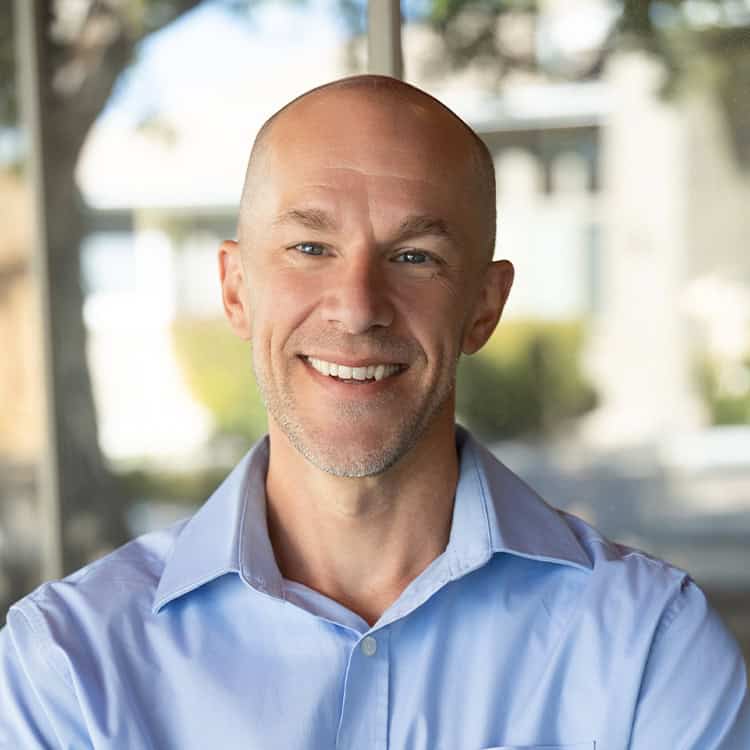 Smiling middle-aged bald man wearing a light-blue button-down shirt, facing the camera and relaxed; standing indoors by a large window with a sunlit, blurred residential background.