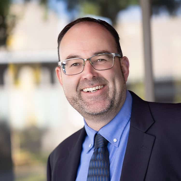 Middle-aged man wearing rimmed glasses and a suit smiles openly, head slightly tilted; short beard, blue shirt and patterned tie provide contrast against a soft-focused outdoor building backdrop.
