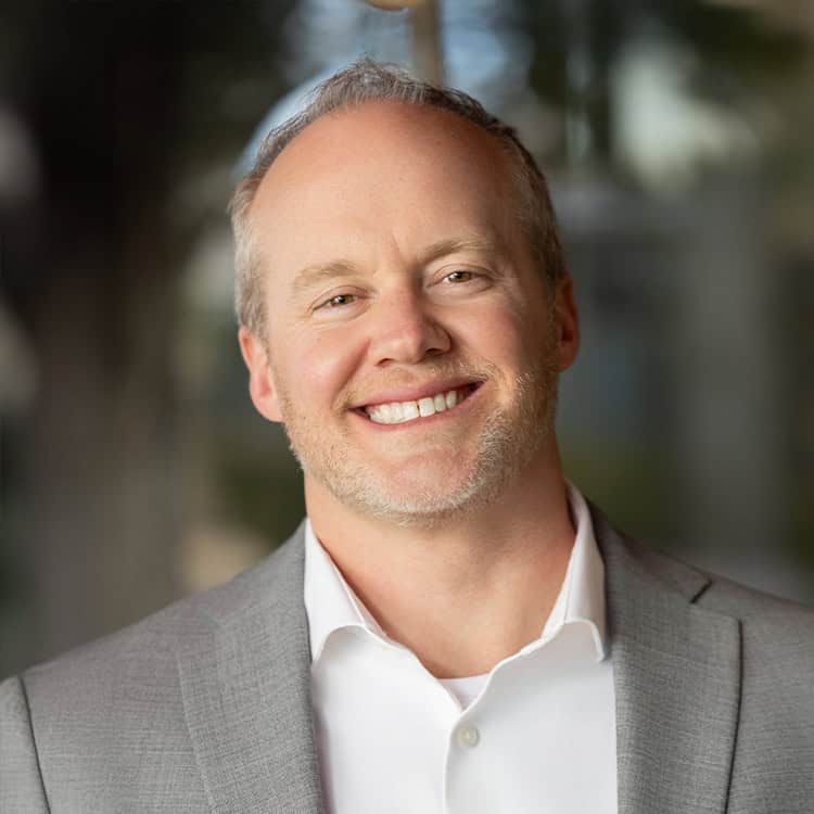 Smiling middle-aged man in a gray suit and white shirt, tilting his head slightly toward the camera, standing outdoors with soft-focus trees and a building in the background.