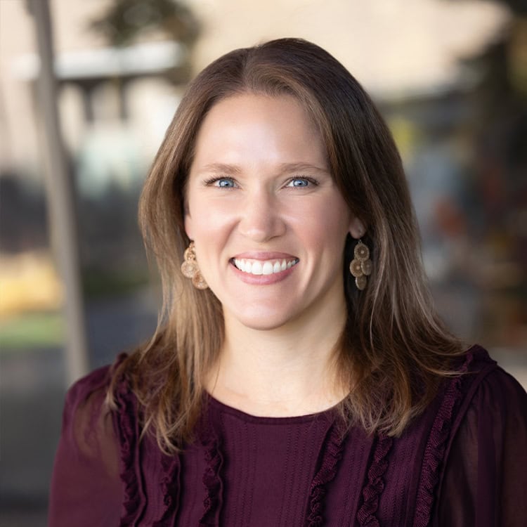 Woman with shoulder-length brown hair smiling at camera, wearing maroon textured blouse and gold dangling earrings, posed against a softly blurred indoor/outdoor background.