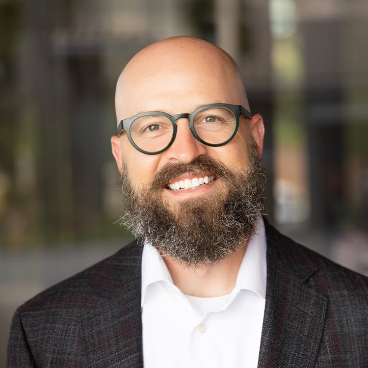 Bald man with round glasses and a full beard — smiling broadly, showing teeth — standing against a softly blurred indoor background, wearing a dark plaid blazer and white dress shirt.