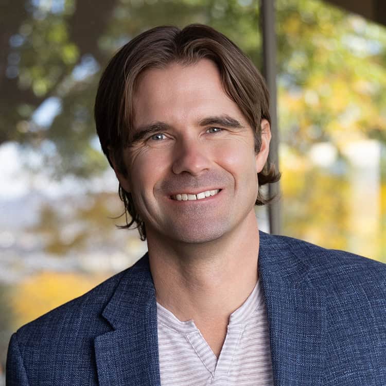 A smiling man with brown hair looks at the camera, wearing a blue blazer and light V-neck shirt, indoors before a large window showing blurred green and yellow foliage.