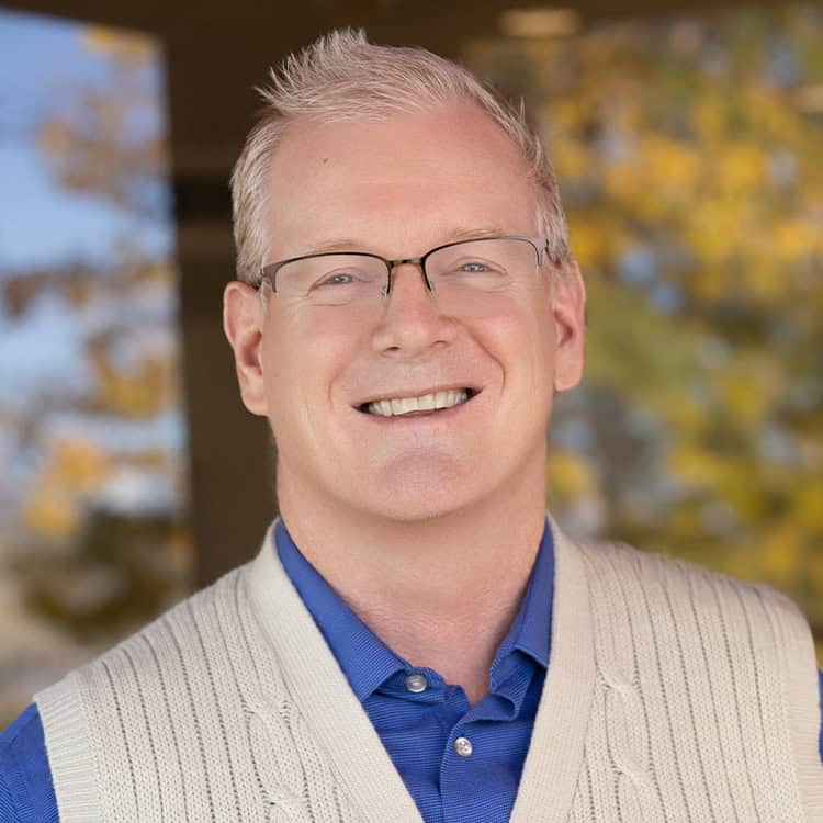 Smiling middle-aged man with short gray hair and glasses smiles at the camera, standing outdoors before blurred autumn foliage, wearing a blue shirt and cream cable-knit vest.