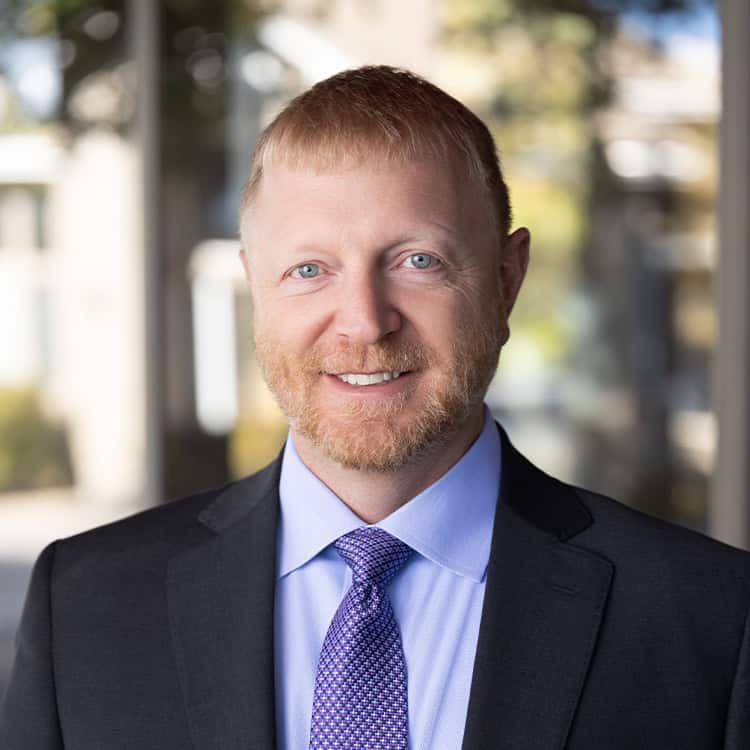 A red-haired bearded man with blue eyes in a dark suit and purple tie smiles directly at the camera, standing against a softly blurred glass building and trees background.