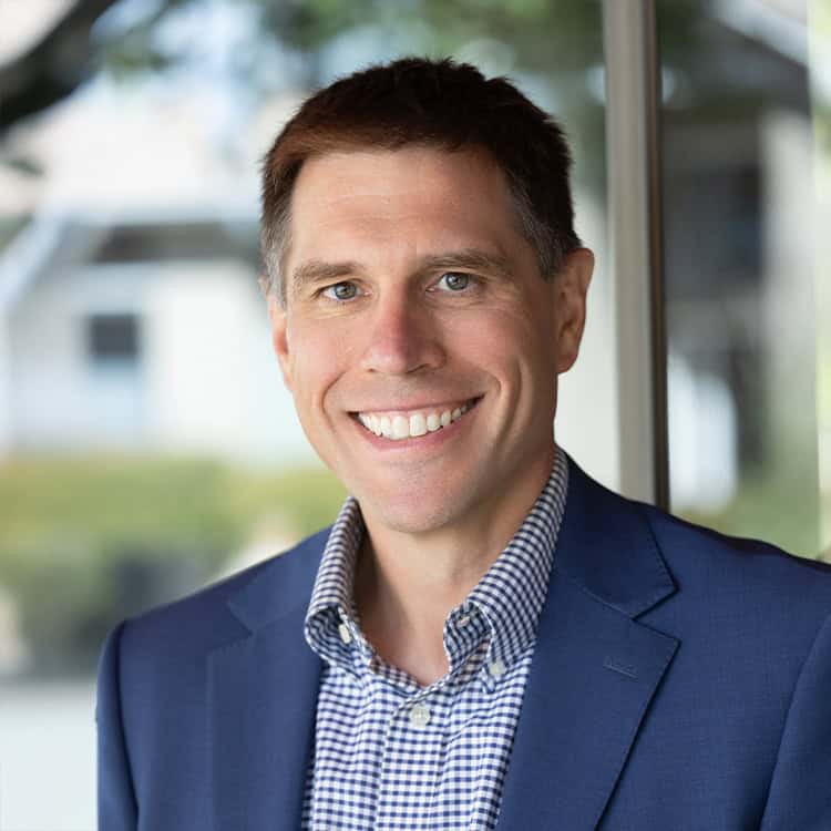 A smiling man looks at the camera, wearing a blue blazer and checked shirt, standing by a glass window with a softly blurred outdoor background.