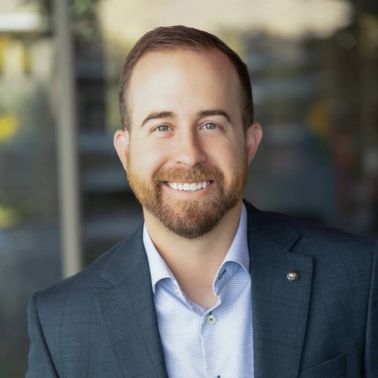 Bearded man smiles directly at the camera (object-action), wearing a light-blue shirt and dark-blue checked blazer (context), standing before a softly blurred modern glass-office interior.