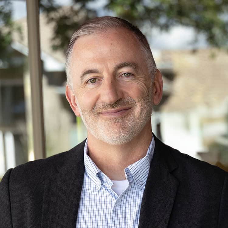 Middle-aged man with short gray hair and trimmed beard, smiling gently at camera, wearing a checked shirt and dark blazer, standing outdoors by a glass window with blurred trees.