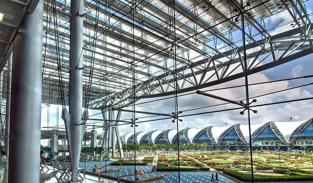 Glass curtain wall frames landscaped courtyard and rows of arched terminal roofs; massive steel columns and trussed roof structure rise inside a spacious, sunlit airport concourse.