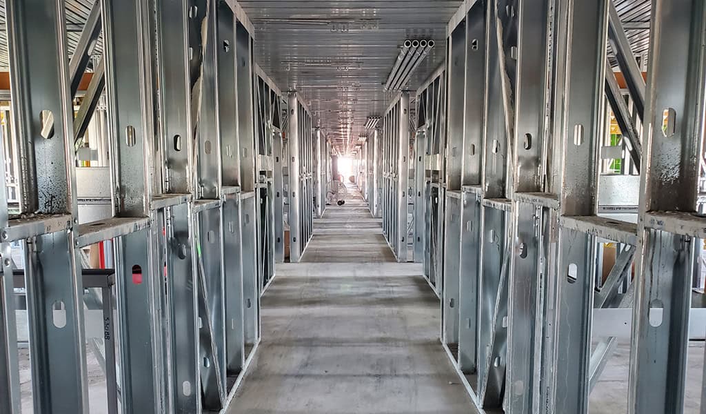 Metal wall studs form a symmetrical corridor leading toward a bright far end, with a concrete floor, overhead decking and exposed pipes inside an unfinished building construction site.