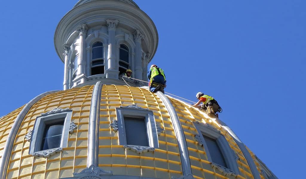 A gilded dome is being repaired by three tethered construction workers in high-visibility vests and helmets, near arched windows beneath a cupola against a clear blue sky.