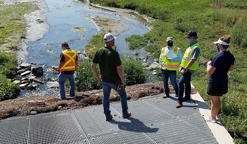 Group of five inspection workers stand on a metal grate and grassy bank, observing a shallow stream and rocky culvert; several wear high-visibility vests, hard hats, and face coverings.