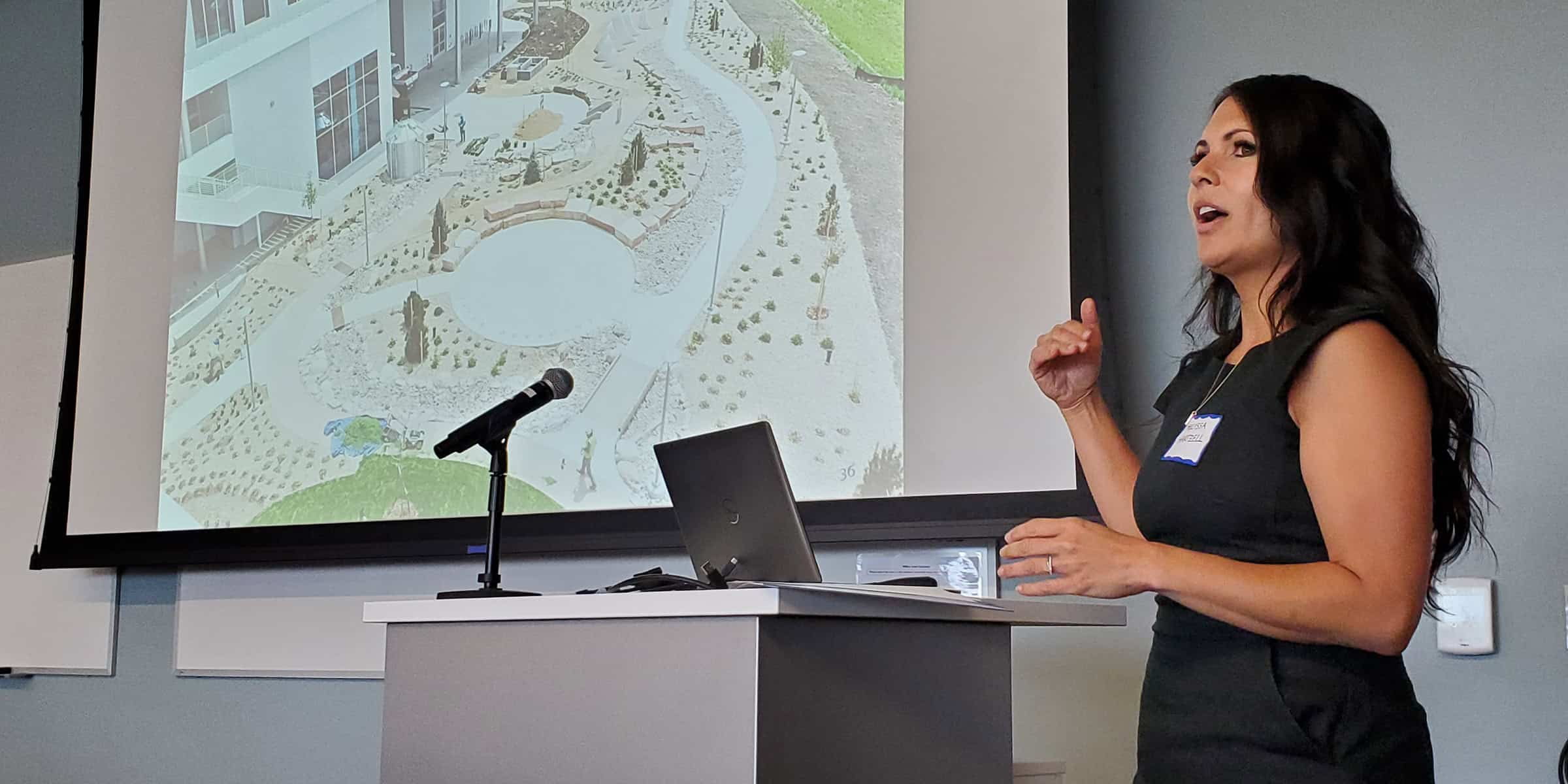 Woman presenter gesturing and speaking beside a projected landscape plan, standing at a podium with microphone and laptop in a small conference room.

Text visible: name tag — handwritten, illegible; projection lower-right — "36".