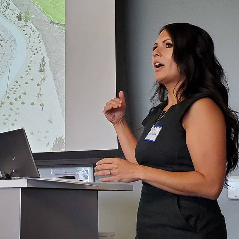 Woman presenting at a podium gestures toward a projected site map, laptop on the lectern, in a conference room. A name tag is visible but I cannot transcribe the person's name.