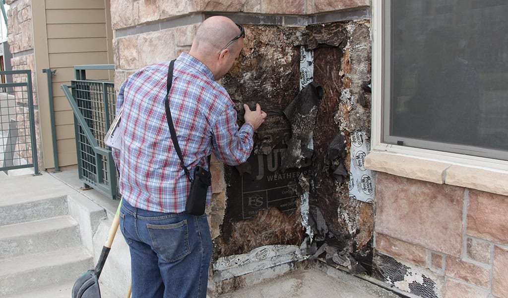 Man peels back charred exterior siding, inspecting ruined sheathing beside a window and concrete steps. Text visible on sheathing: "JUTA WEATHERBARRIER". Repeated on flashing tape: "TYVEK".