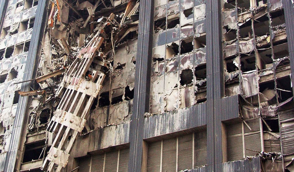 High-rise facade with a collapsed multi-story steel structure hanging diagonally, floors exposed and windows blown out, scorched surfaces and debris strewn across the exterior in an urban setting.