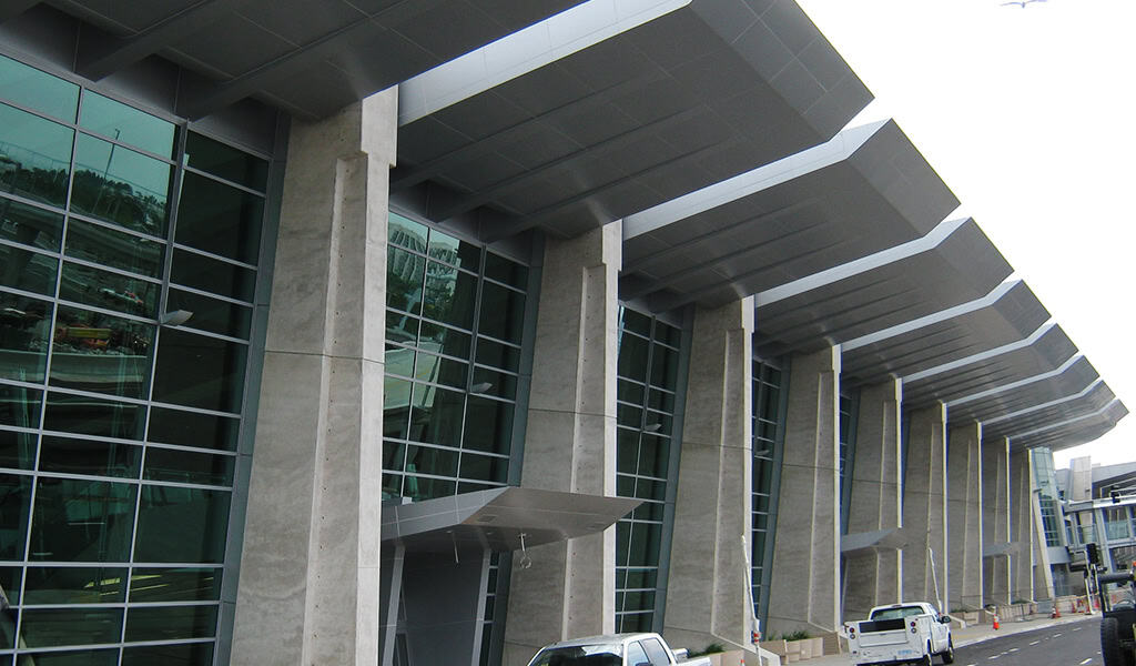 Modern glass-and-concrete façade features tall concrete columns supporting a stepped metal canopy, lining a curb with parked trucks and a roadway under an overcast sky.