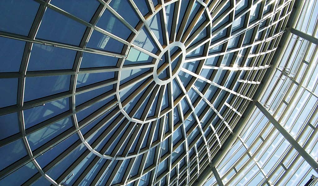 Glass dome skylight featuring radial steel ribs converging at a central ring, casting geometric shadows across blue-tinted glass panels within a modern atrium.