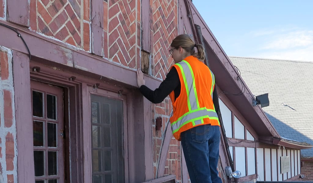 Worker in an orange reflective vest touches and inspects a wooden beam while standing on a ladder against a brick-and-tudor façade of a multi-story building under clear sky.