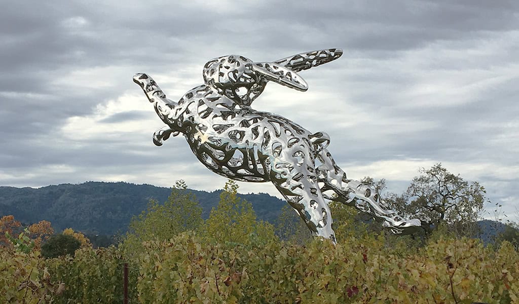 Large polished-metal hare sculpture leaps forward, body perforated with organic cutouts; suspended above vineyard foliage, with rolling wooded hills and a cloudy, overcast sky in the background.
