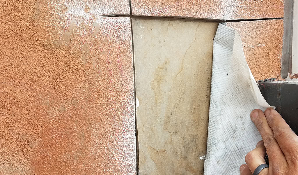 White membrane being peeled back by a hand to reveal a beige marble tile set into a cracked orange stucco wall beside a black metal ledge.