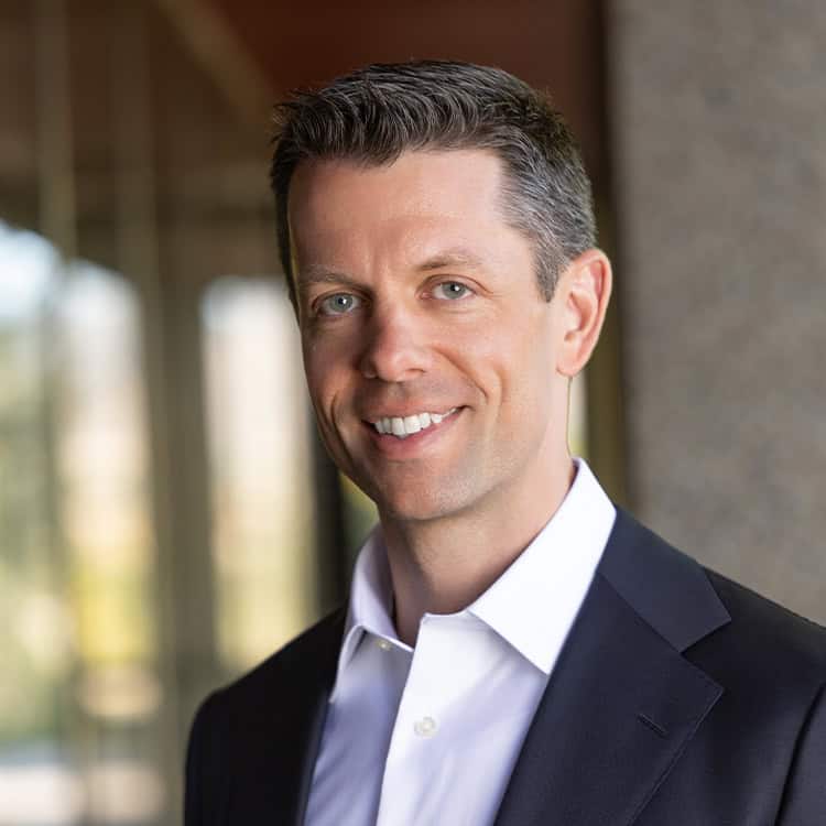 Smiling man in a dark suit and white shirt, facing the camera; standing near a stone column with a blurred glass-windowed office background, warm natural lighting.
