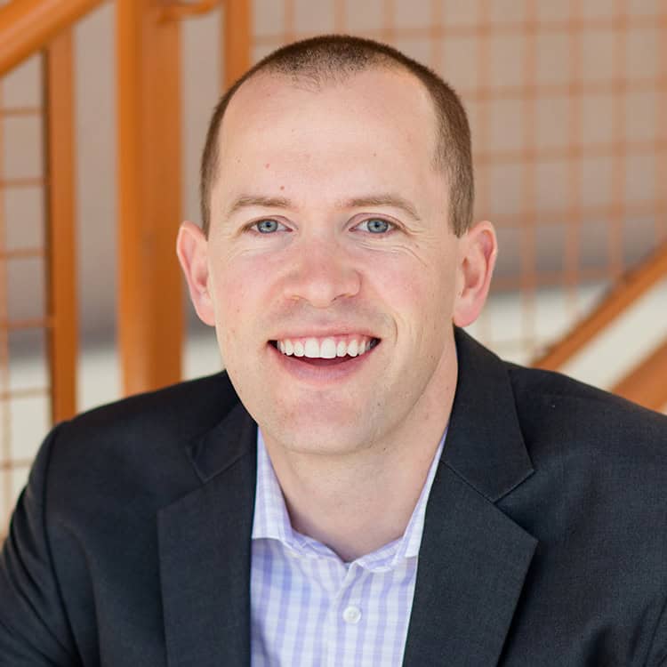 Man (close-up headshot) smiling at camera, wearing a dark suit jacket and light checkered shirt, seated in front of an orange metal staircase with a softly blurred background.